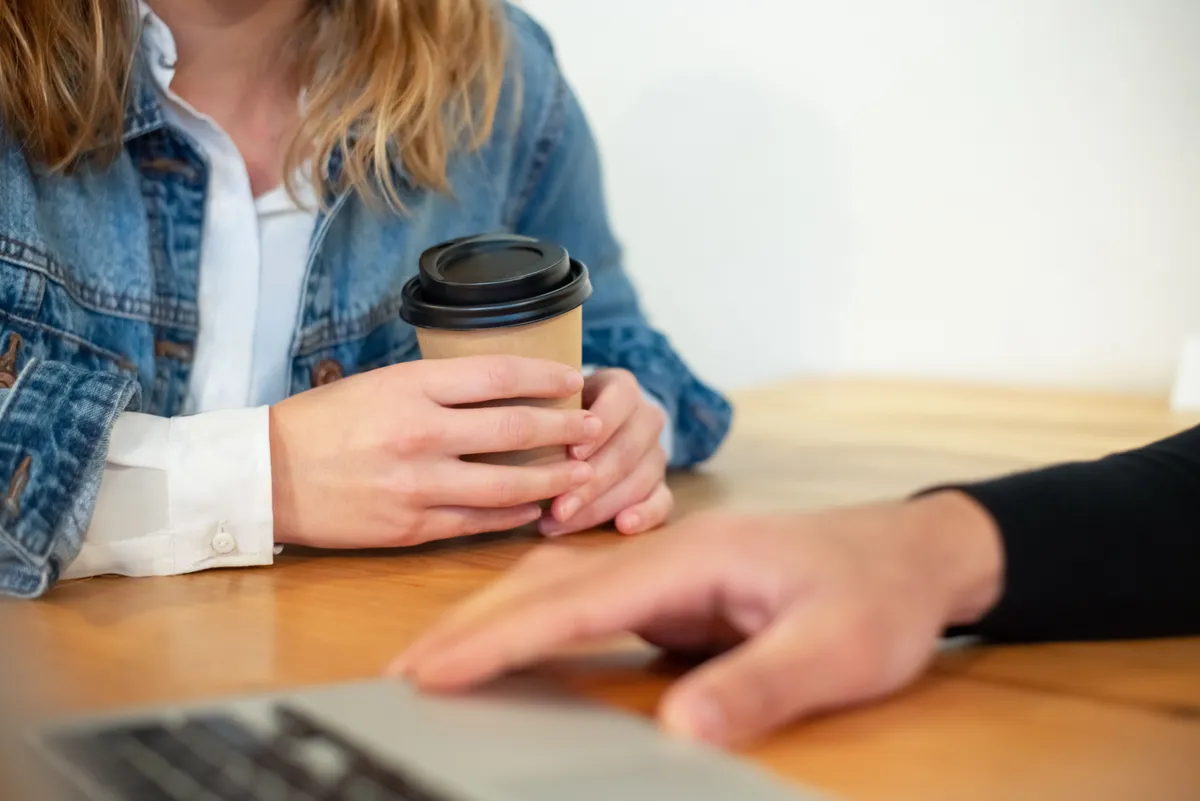 Mulher segurando um copo de café em uma mesa, conversando com outra pessoa, cenário de uma conversa tranquila sobre expectativas
