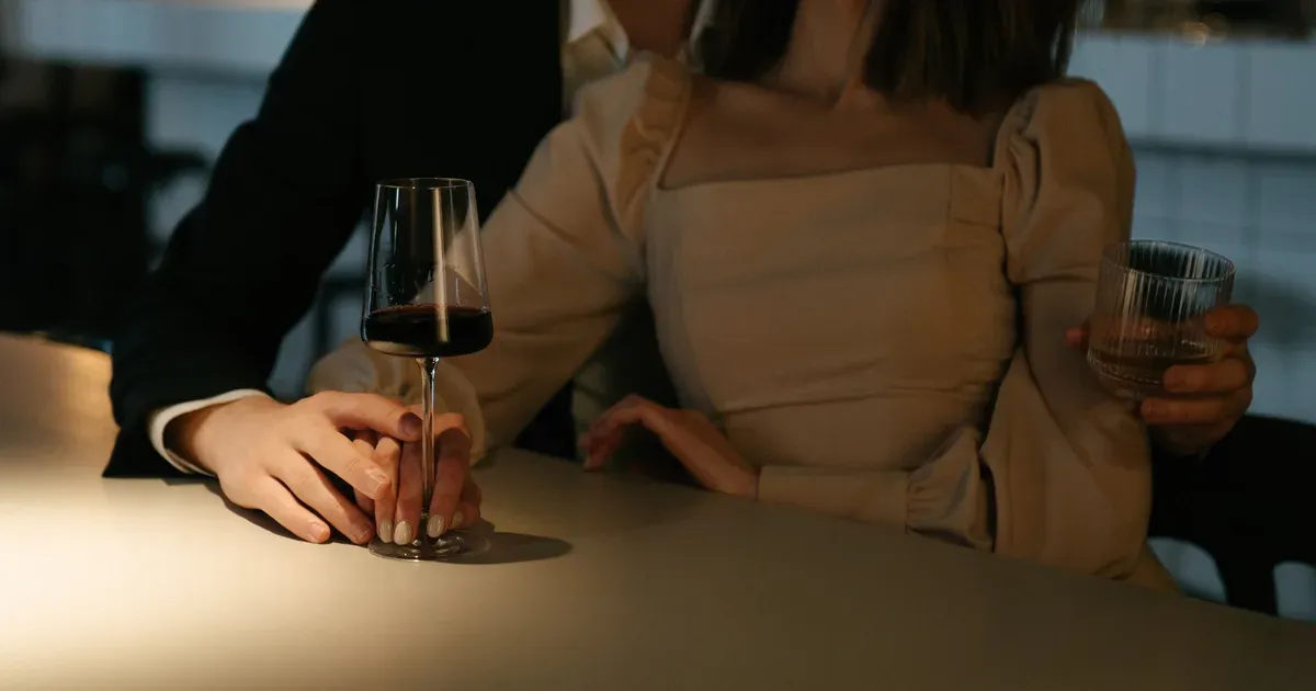 Casal elegante num bar, ele de terno com a mão sobre a dela segurando uma taça de vinho, ilustrando o post sobre hipergamia