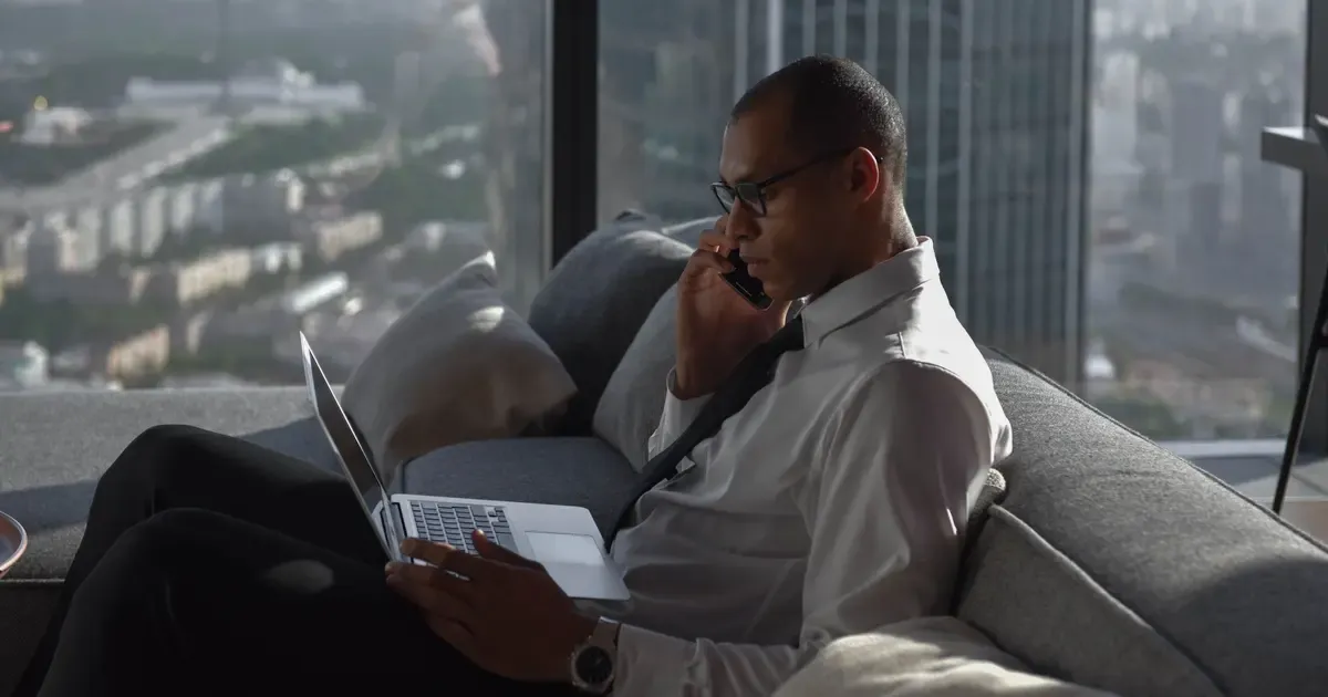 Empresário de camisa social falando ao telefone com o notebook no colo, num apartamento com vista para a cidade, ilustrando o post sobre namorar empresário