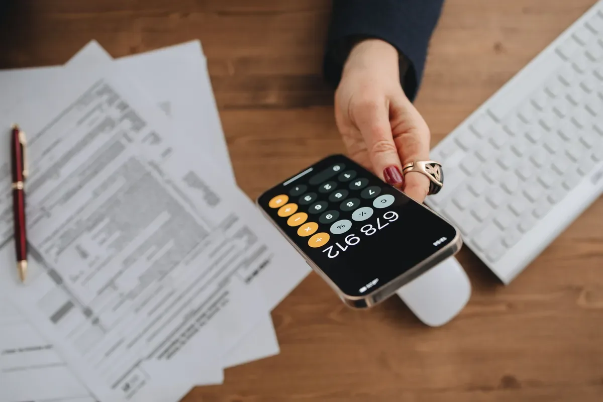 Mão segurando celular com calculadora sobre uma mesa com papéis, ilustrando o cálculo de valores em relacionamento sugar